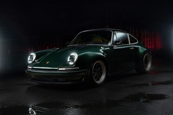 A dark green classic Porsche sports car is parked on a wet surface at night, with its headlights on and a dark, blurred background illuminated by subtle red and white lighting.