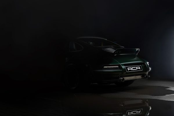 A dark, moody photo of a green sports car parked on a reflective surface, with only the rear of the car illuminated. The car features a spoiler and a license plate reading ACA. The background fades into blackness.