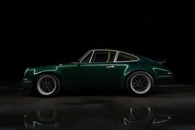 A dark green classic sports car is shown in profile on a wet surface, reflecting light. The background is black, highlighting the car’s sleek lines and vintage design.